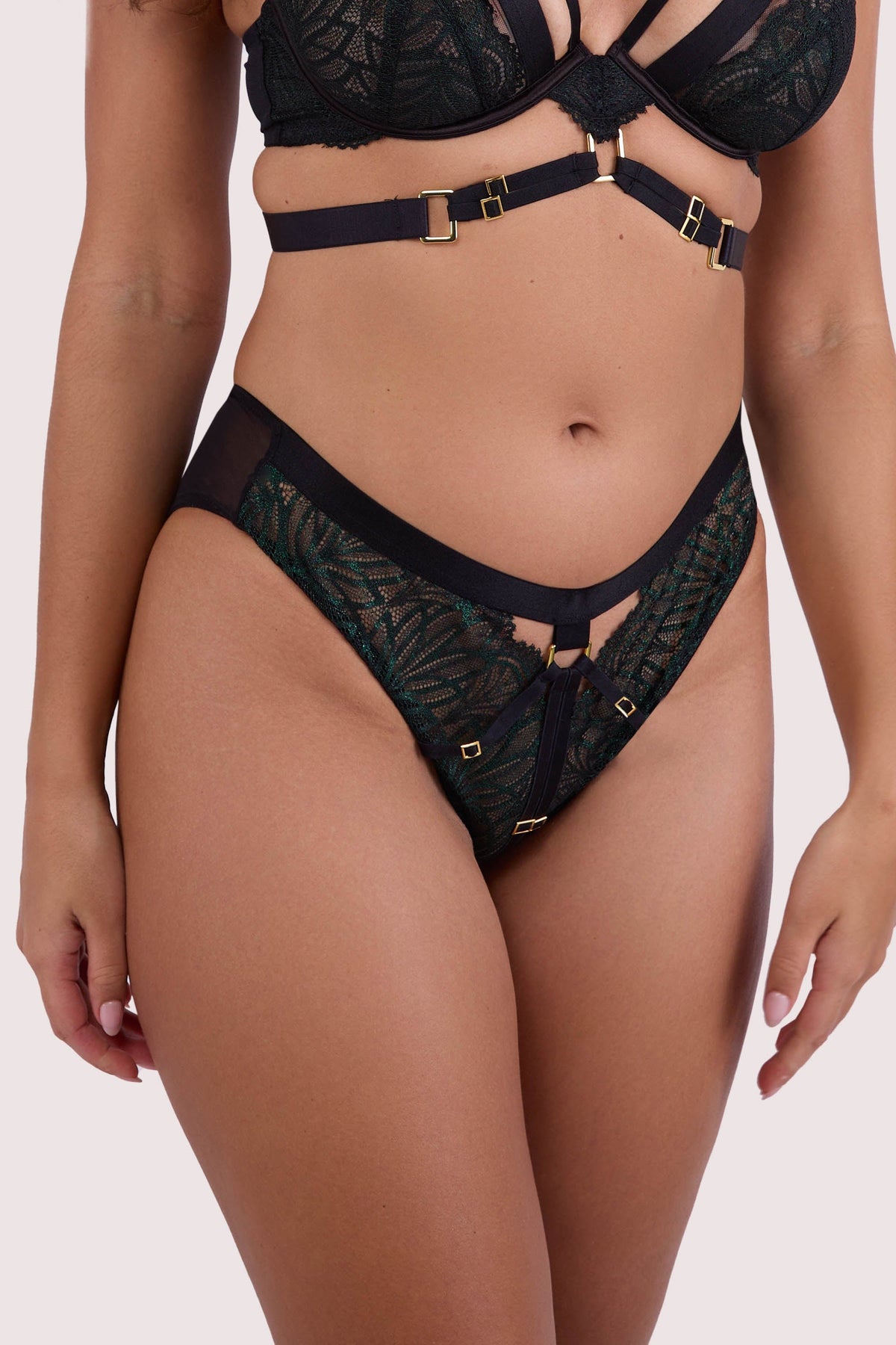 Model wears dark green wet look lace thong with black harness straps and gold-tone hardware