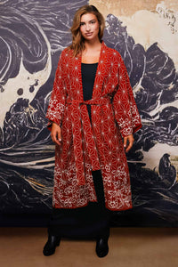 Model wears belted terracotta kimono style robe with ivory embroidery over long black dress