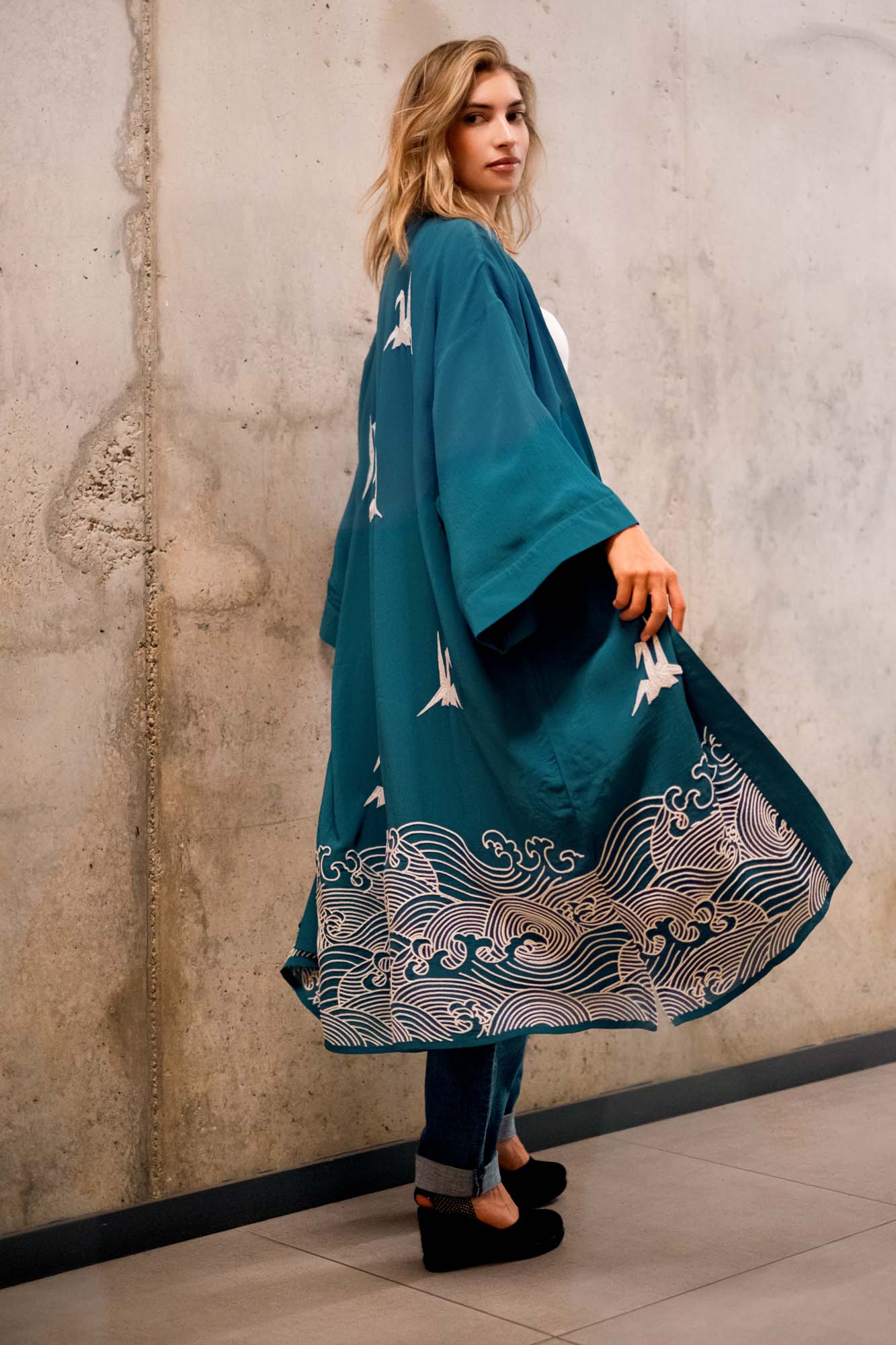 Model shows back of teal kimono style robe embroidered with ivory herons and Japanese waves