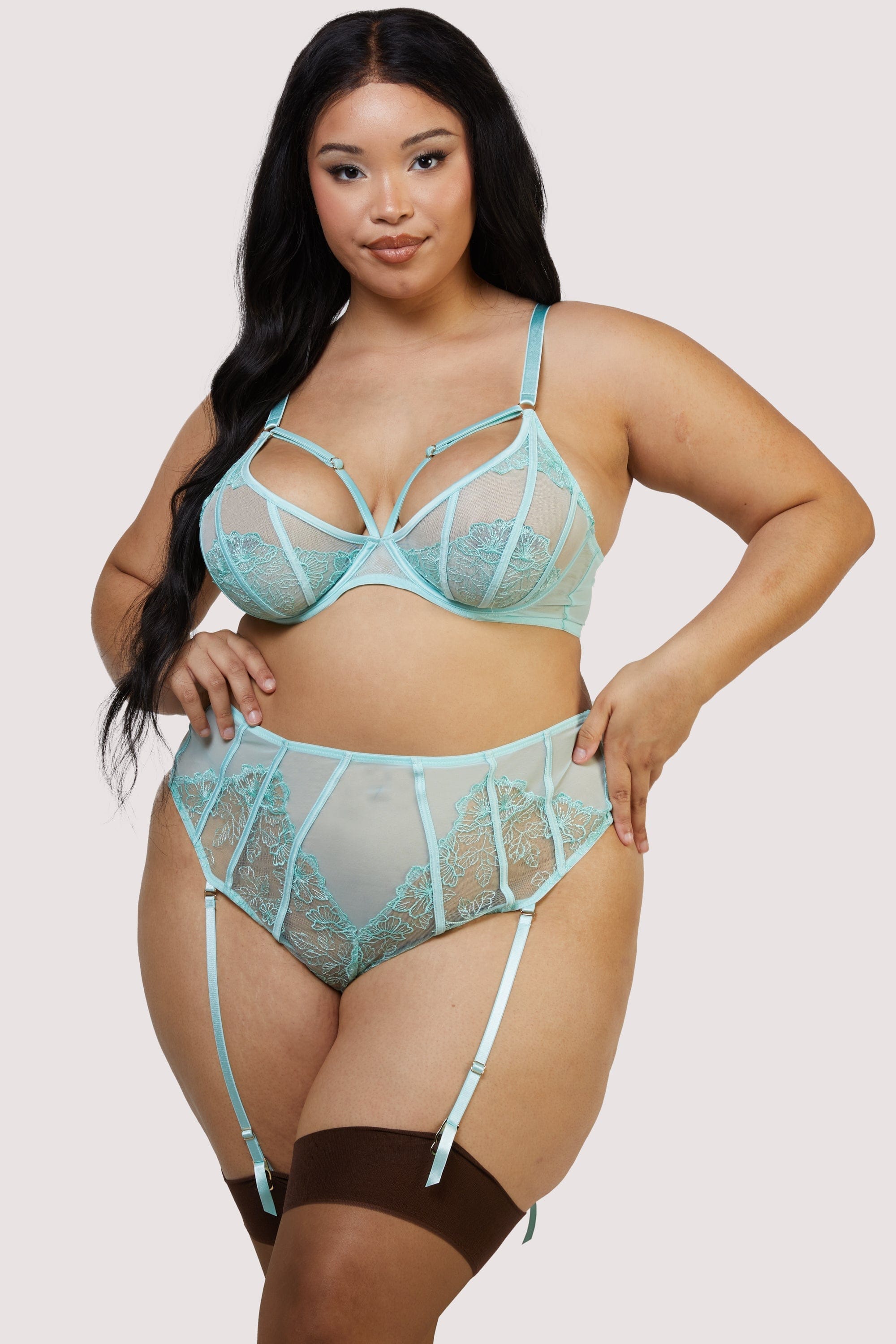 Model wears aqua lingerie set featuring sheer plunge bra with floral embroidery and matching high waist suspender thong with satin straps, paired with dark brown thigh-high stockings