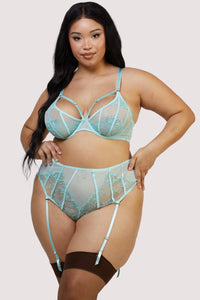 Model wears aqua lingerie set featuring sheer plunge bra with floral embroidery and matching high waist suspender thong with satin straps, paired with dark brown thigh-high stockings