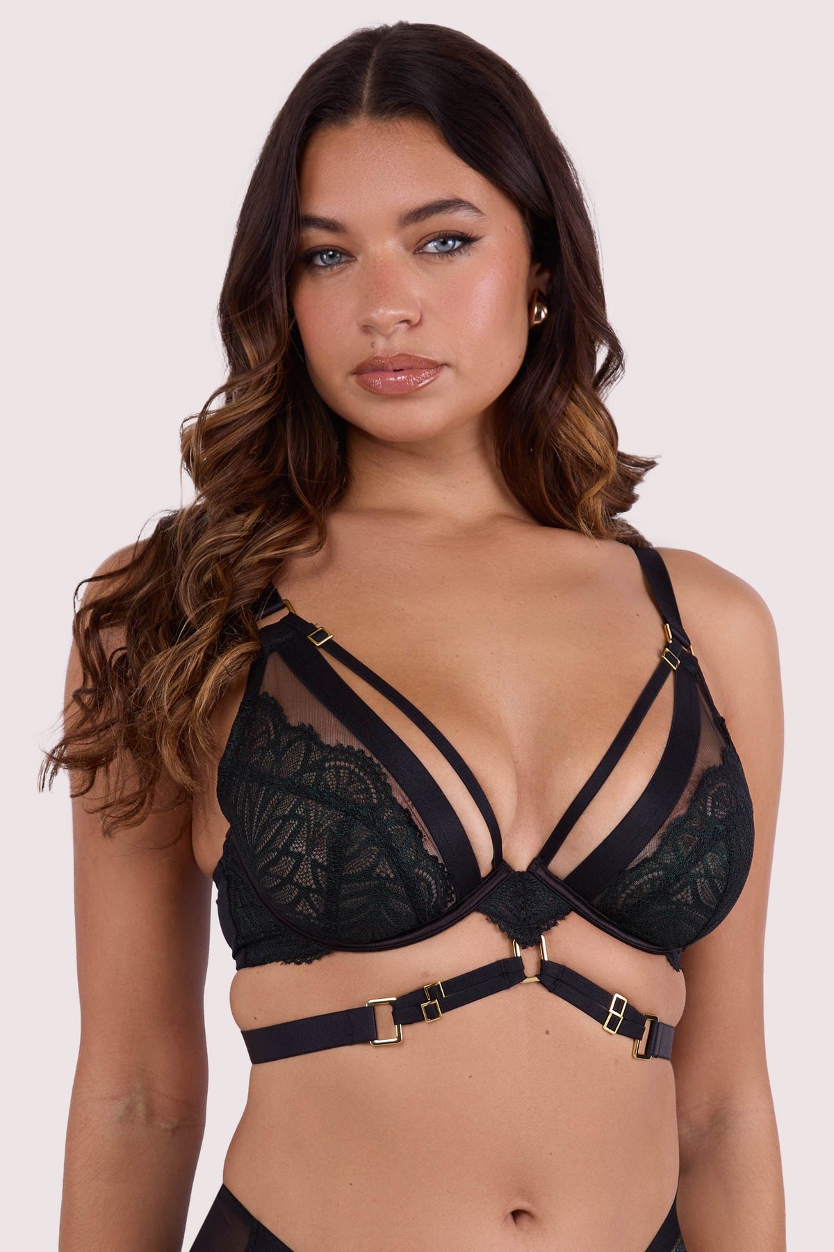 Model wears dark green wet-look  plunge bra with  underwire cups, strappy harness design, and gold buckle detailing